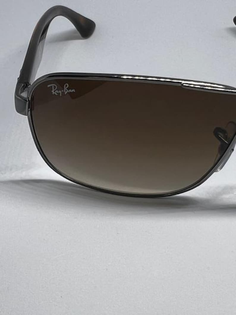 Pode incluir: &Oacute;culos de sol Ray-Ban com arma&ccedil;&atilde;o met&aacute;lica escura e lentes degrad&ecirc; castanhas. A lente esquerda apresenta o log&oacute;tipo branco Ray-Ban no canto superior. As hastes t&ecirc;m um padr&atilde;o de tartaruga escuro e a ponte tem um acabamento met&aacute;lico.
