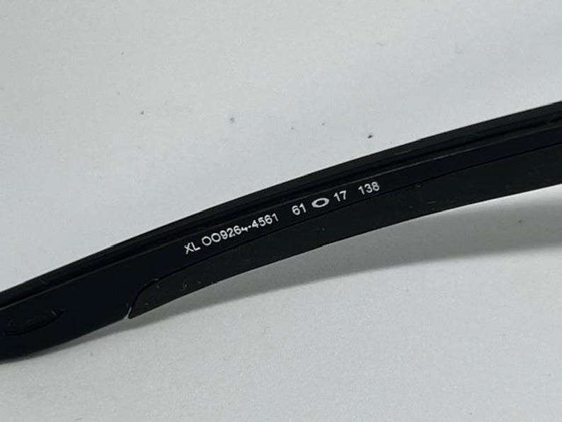 May include: A black sunglass temple arm featuring white printed text on the inner side. The text reads XL OO9264-4561 61 17 138, indicating the model number and specific frame measurements in millimeters for this eyewear component.
