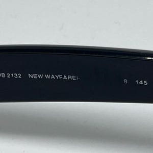 Puede incluir: El interior de una patilla de gafas de sol negra con texto impreso en blanco que dice RB 2132 NEW WAYFARER 8 145 3N. La superficie es lisa y oscura, mostrando la informaci&oacute;n del modelo claramente sobre el material pl&aacute;stico negro.