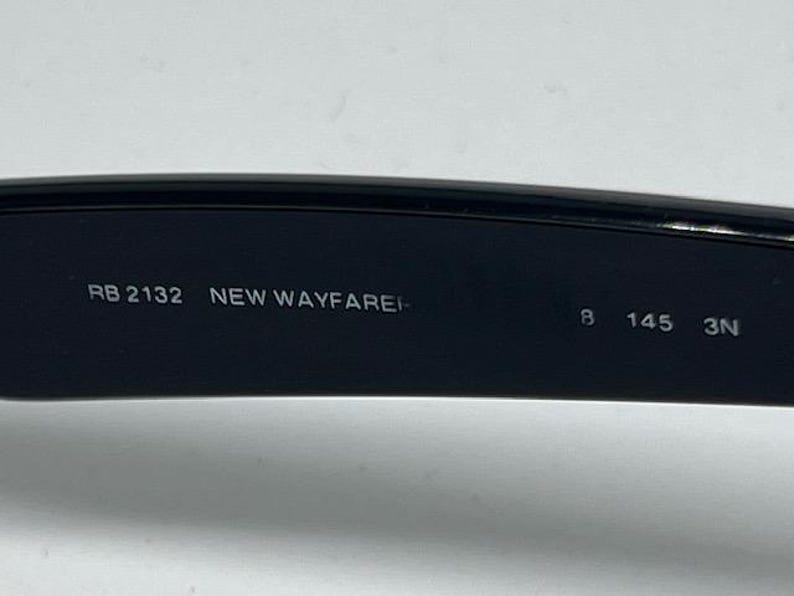 Puede incluir: El interior de una patilla de gafas de sol negra con texto impreso en blanco que dice RB 2132 NEW WAYFARER 8 145 3N. La superficie es lisa y oscura, mostrando la informaci&oacute;n del modelo claramente sobre el material pl&aacute;stico negro.