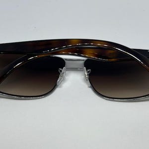 Pode incluir: &Oacute;culos de sol Ray-Ban com arma&ccedil;&atilde;o de metal prateado e lentes retangulares castanhas degrad&ecirc;. As hastes s&atilde;o feitas de acetato com padr&atilde;o de tartaruga castanho escuro e exibem o log&oacute;tipo Ray-Ban em letras brancas em ambos os lados.