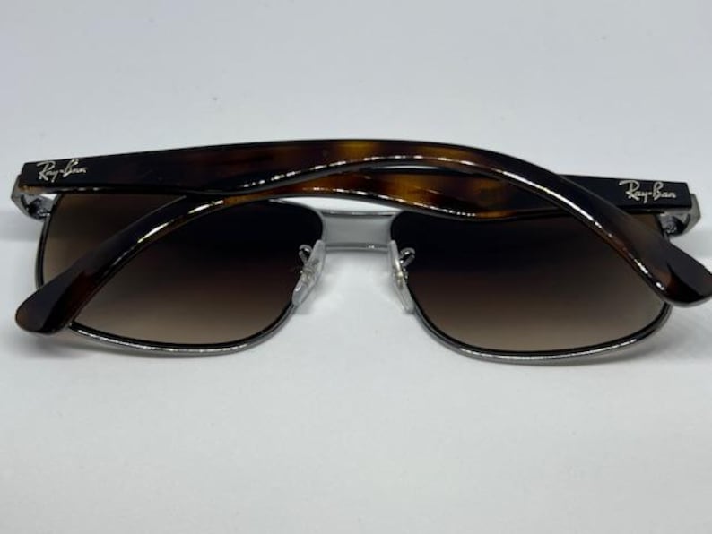 Pode incluir: &Oacute;culos de sol Ray-Ban com arma&ccedil;&atilde;o de metal prateado e lentes retangulares castanhas degrad&ecirc;. As hastes s&atilde;o feitas de acetato com padr&atilde;o de tartaruga castanho escuro e exibem o log&oacute;tipo Ray-Ban em letras brancas em ambos os lados.
