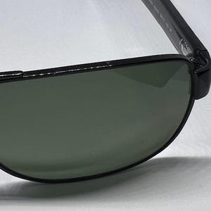 May include: Black aviator-style sunglasses featuring dark green tinted lenses and a thin black metal frame. The temple arm displays white printed text detailing the model specifications, including the numbers 5519 001 145 3P.