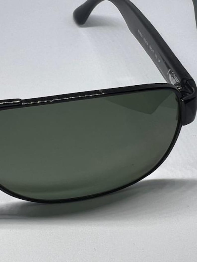 May include: Black aviator-style sunglasses featuring dark green tinted lenses and a thin black metal frame. The temple arm displays white printed text detailing the model specifications, including the numbers 5519 001 145 3P.