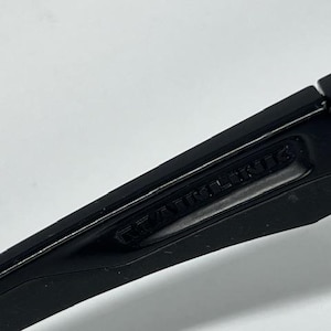 May include: A matte black plastic component with a hinged section and a recessed area featuring the embossed text "RAWLINGS". The object has a smooth, angular design with a visible pivot point and a clean, uniform dark finish throughout the piece.