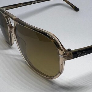 May include: Aviator sunglasses featuring a translucent beige and brown tortoiseshell frame with a double bridge design. The lenses are tinted brown, and the temples display the Ray-Ban logo in white lettering on a black background.