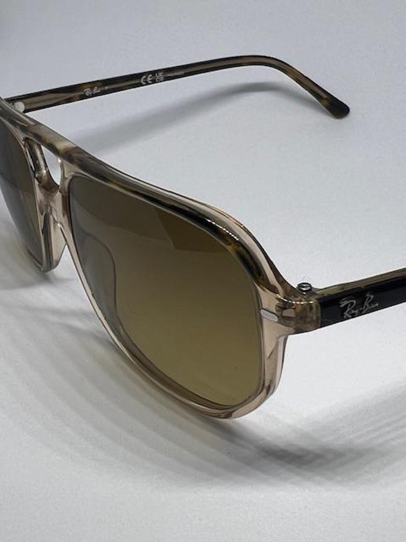 May include: Aviator sunglasses featuring a translucent beige and brown tortoiseshell frame with a double bridge design. The lenses are tinted brown, and the temples display the Ray-Ban logo in white lettering on a black background.