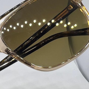 May include: Sunglasses with a translucent champagne-coloured frame and dark brown lenses. The inner temple arm features the printed text RB 220, THE MARSHAL ONE, 1292/M2, and 54-19. The lenses have a reflective surface showing a series of small light dots.