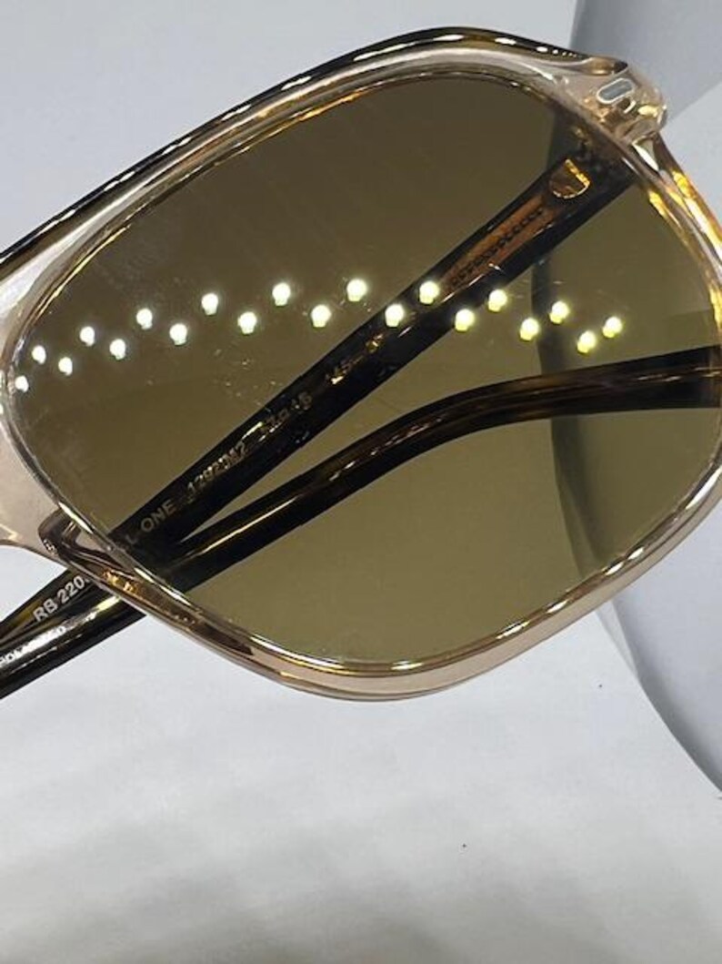 May include: Sunglasses with a translucent champagne-coloured frame and dark brown lenses. The inner temple arm features the printed text RB 220, THE MARSHAL ONE, 1292/M2, and 54-19. The lenses have a reflective surface showing a series of small light dots.