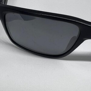May include: A pair of black sunglasses featuring a thick, solid black frame and dark, tinted lenses. The lenses exhibit visible surface scratches across the center, while the frame maintains a smooth, matte black finish throughout the structure.