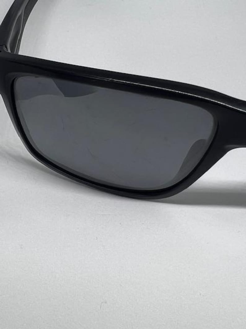 May include: A pair of black sunglasses featuring a thick, solid black frame and dark, tinted lenses. The lenses exhibit visible surface scratches across the center, while the frame maintains a smooth, matte black finish throughout the structure.