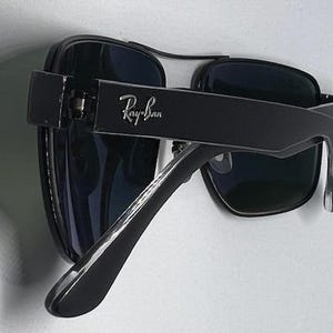May include: Black sunglasses with dark tinted lenses and a thick frame. The Ray-Ban logo is printed in silver on the outer side of the temple arm. The design features a double bridge across the top of the lenses and curved temple tips for a secure fit.