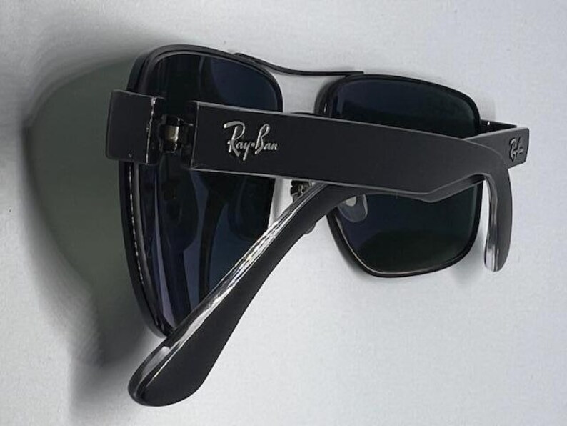 May include: Black sunglasses with dark tinted lenses and a thick frame. The Ray-Ban logo is printed in silver on the outer side of the temple arm. The design features a double bridge across the top of the lenses and curved temple tips for a secure fit.