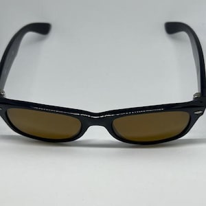Puede incluir: Gafas de sol con montura negra de forma rectangular cl&aacute;sica y bordes redondeados. Las lentes tienen un tono marr&oacute;n s&oacute;lido y la montura incluye peque&ntilde;os detalles met&aacute;licos de color plateado en las esquinas exteriores de las varillas.