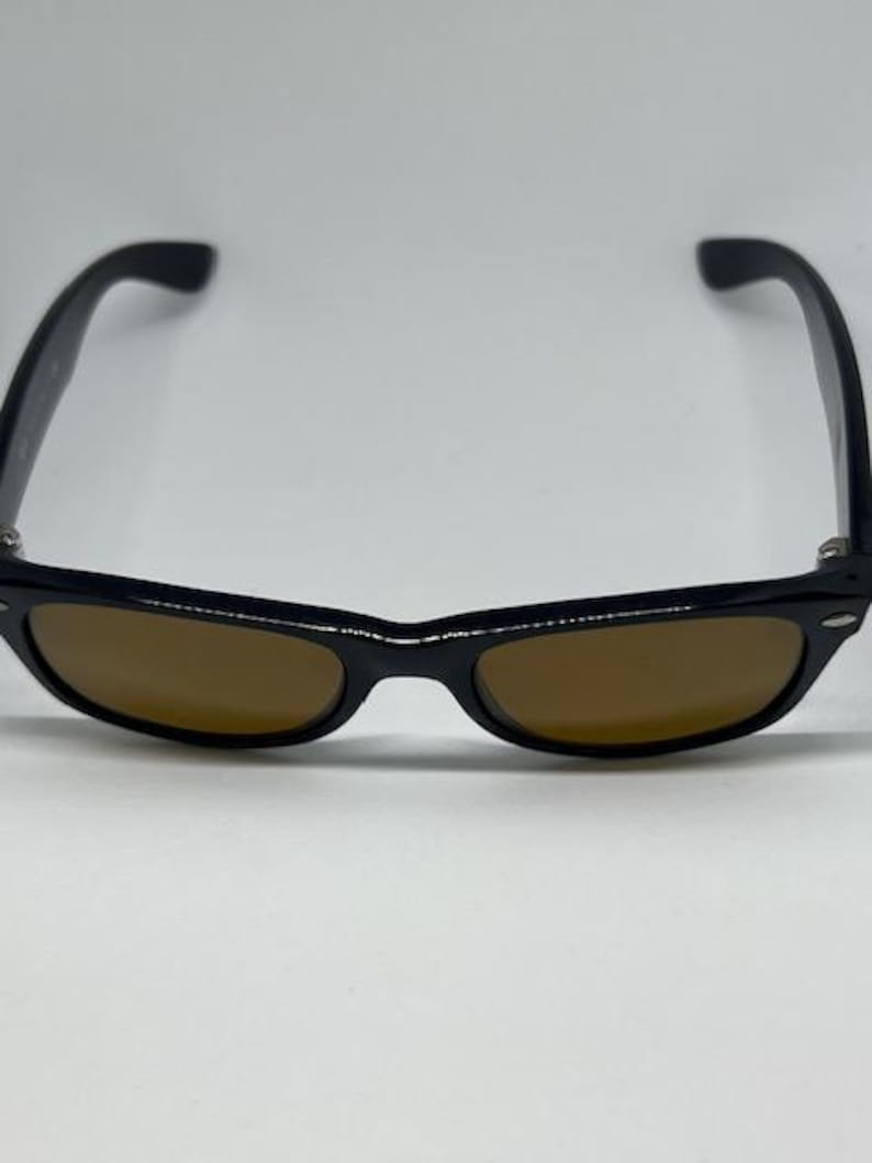 Puede incluir: Gafas de sol con montura negra de forma rectangular cl&aacute;sica y bordes redondeados. Las lentes tienen un tono marr&oacute;n s&oacute;lido y la montura incluye peque&ntilde;os detalles met&aacute;licos de color plateado en las esquinas exteriores de las varillas.