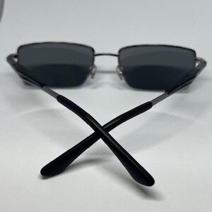 Puede incluir: Gafas de sol rectangulares con lentes de color gris oscuro y una montura fina de metal plateado. Las varillas est&aacute;n construidas con una combinaci&oacute;n de metal plateado y puntas de pl&aacute;stico negro que se cruzan en el centro de la montura.