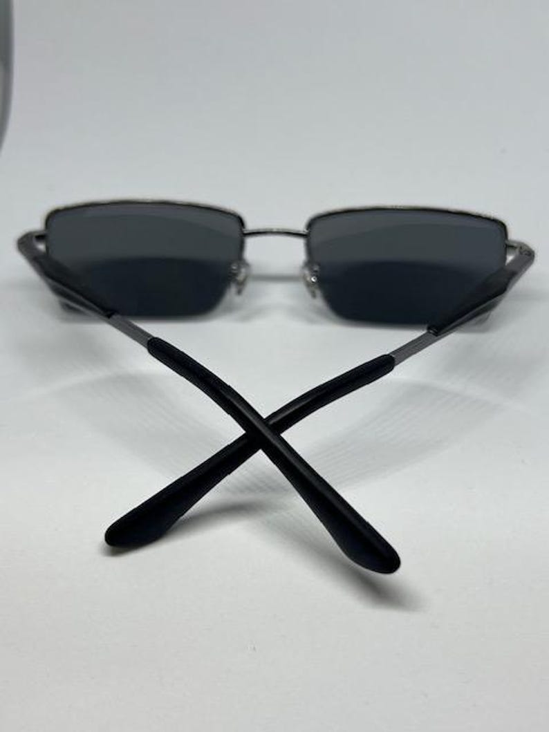 Puede incluir: Gafas de sol rectangulares con lentes de color gris oscuro y una montura fina de metal plateado. Las varillas est&aacute;n construidas con una combinaci&oacute;n de metal plateado y puntas de pl&aacute;stico negro que se cruzan en el centro de la montura.