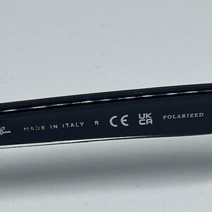 May include: The inner temple of a black pair of sunglasses featuring the white printed text Ray-Ban, MADE IN ITALY, R, the CE mark, the UKCA mark, and the word POLARIZED. The surface is smooth, dark, and glossy with clear, legible white lettering.