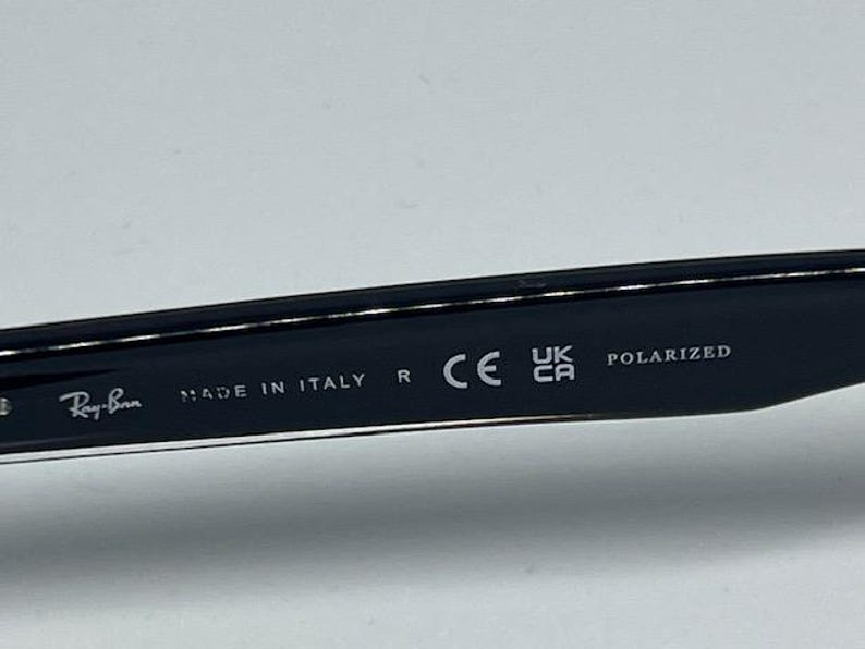 May include: The inner temple of a black pair of sunglasses featuring the white printed text Ray-Ban, MADE IN ITALY, R, the CE mark, the UKCA mark, and the word POLARIZED. The surface is smooth, dark, and glossy with clear, legible white lettering.