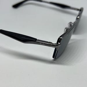 Puede incluir: Unas gafas de sol de metal gris oscuro con puntas de patilla negras. La montura presenta una forma rectangular con lentes oscuras. El logotipo de Ray-Ban est&aacute; impreso en blanco en el lateral de la patilla met&aacute;lica cerca de la bisagra.