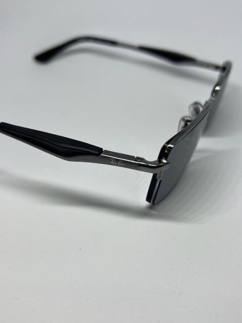Puede incluir: Unas gafas de sol de metal gris oscuro con puntas de patilla negras. La montura presenta una forma rectangular con lentes oscuras. El logotipo de Ray-Ban est&aacute; impreso en blanco en el lateral de la patilla met&aacute;lica cerca de la bisagra.