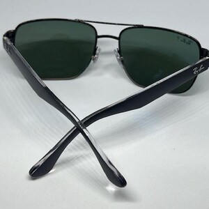May include: Black aviator sunglasses with dark green polarized lenses. The thin metal frame features a double bridge design and adjustable nose pads. The temples are black with a clear tip, and the Ray-Ban logo is printed on the right lens and temple.