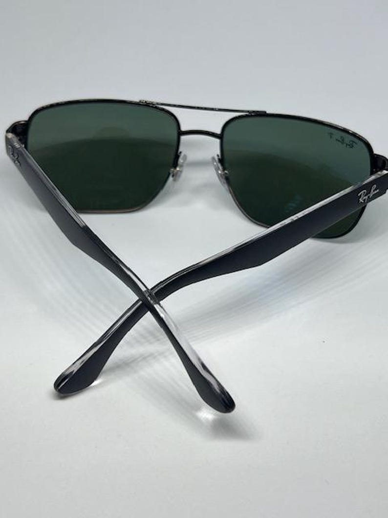 May include: Black aviator sunglasses with dark green polarized lenses. The thin metal frame features a double bridge design and adjustable nose pads. The temples are black with a clear tip, and the Ray-Ban logo is printed on the right lens and temple.