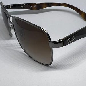 Pode incluir: &Oacute;culos de sol Ray-Ban com arma&ccedil;&atilde;o de metal cinza-chumbo, design de ponte dupla e lentes degrad&ecirc; marrons. As hastes s&atilde;o feitas de material com padr&atilde;o de tartaruga marrom com o logotipo Ray-Ban impresso em prata nas dobradi&ccedil;as laterais.