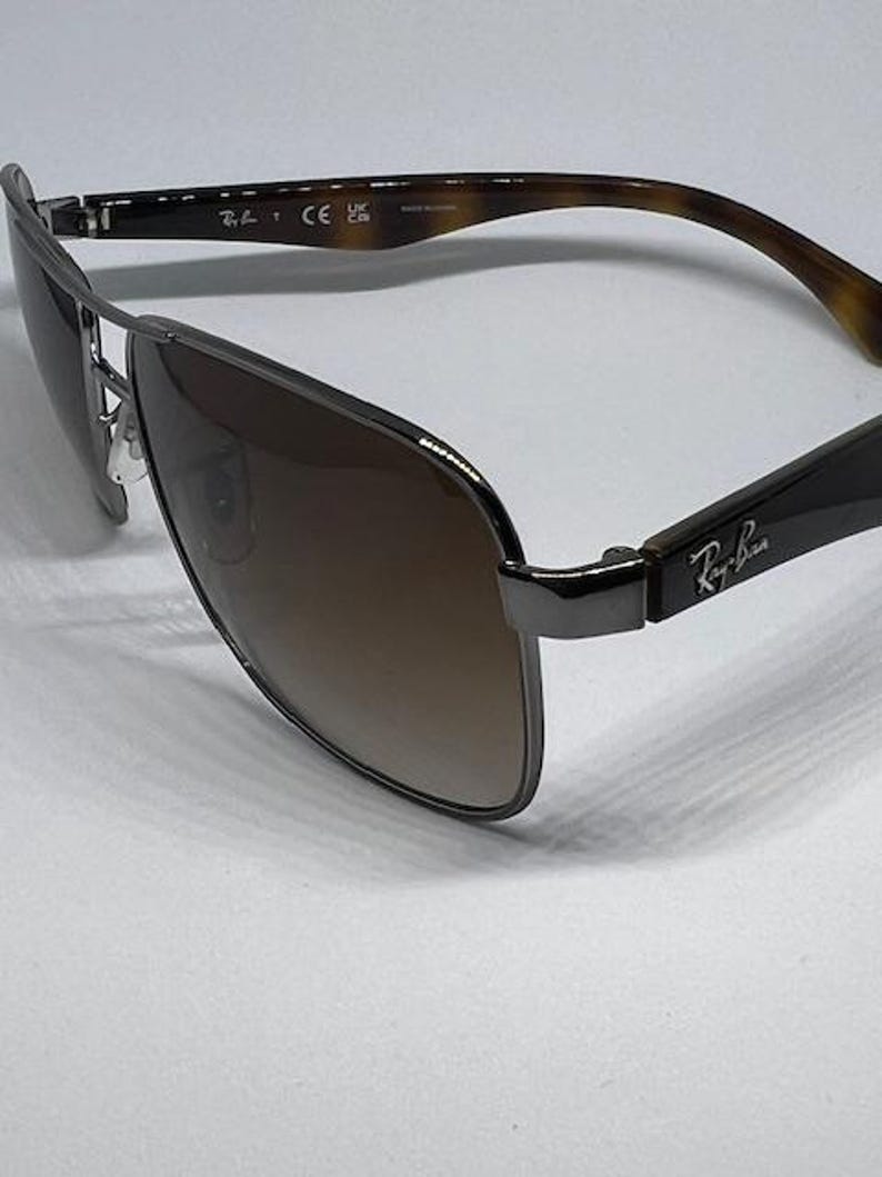 Pode incluir: &Oacute;culos de sol Ray-Ban com arma&ccedil;&atilde;o de metal cinza-chumbo, design de ponte dupla e lentes degrad&ecirc; marrons. As hastes s&atilde;o feitas de material com padr&atilde;o de tartaruga marrom com o logotipo Ray-Ban impresso em prata nas dobradi&ccedil;as laterais.