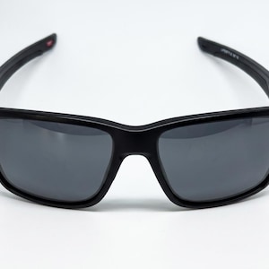 May include: Rectangular sunglasses with a matte black frame and dark gray tinted lenses. The temples are curved and feature a small red accent on the inner side near the hinge, with white text printed on the inner temple arm.