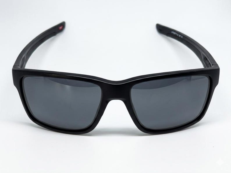 May include: Rectangular sunglasses with a matte black frame and dark gray tinted lenses. The temples are curved and feature a small red accent on the inner side near the hinge, with white text printed on the inner temple arm.