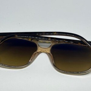 May include: Ray-Ban sunglasses featuring a translucent beige frame with a brown gradient lens. The temples are a dark brown tortoiseshell pattern with the Ray-Ban logo printed in white on the outer side of each temple near the hinge.