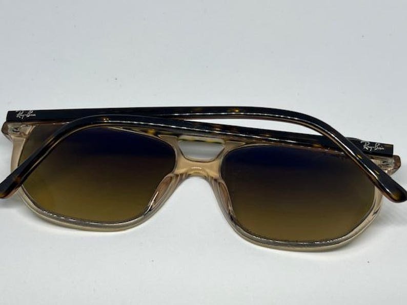 May include: Ray-Ban sunglasses featuring a translucent beige frame with a brown gradient lens. The temples are a dark brown tortoiseshell pattern with the Ray-Ban logo printed in white on the outer side of each temple near the hinge.