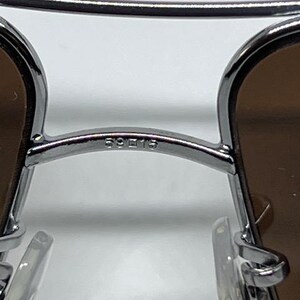 Pode incluir: Um par de &oacute;culos de sol de metal prateado com design de ponte dupla, lentes castanhas e almofadas nasais transparentes. A ponte tem gravado o texto 59 15, indicando a largura da lente e da ponte em mil&iacute;metros.