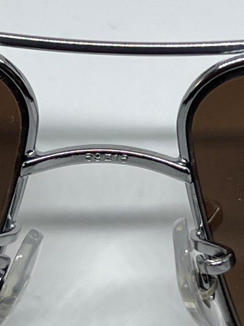 Pode incluir: Um par de &oacute;culos de sol de metal prateado com design de ponte dupla, lentes castanhas e almofadas nasais transparentes. A ponte tem gravado o texto 59 15, indicando a largura da lente e da ponte em mil&iacute;metros.