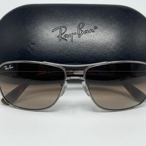 Pode incluir: &Oacute;culos de sol com arma&ccedil;&atilde;o de metal prateado, lentes castanhas degrad&ecirc; e o log&oacute;tipo Ray-Ban na lente superior esquerda. Os &oacute;culos est&atilde;o posicionados &agrave; frente de um estojo de prote&ccedil;&atilde;o r&iacute;gido preto, texturado e oval com o log&oacute;tipo Ray-Ban.