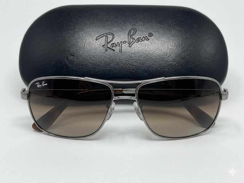 Pode incluir: &Oacute;culos de sol com arma&ccedil;&atilde;o de metal prateado, lentes castanhas degrad&ecirc; e o log&oacute;tipo Ray-Ban na lente superior esquerda. Os &oacute;culos est&atilde;o posicionados &agrave; frente de um estojo de prote&ccedil;&atilde;o r&iacute;gido preto, texturado e oval com o log&oacute;tipo Ray-Ban.