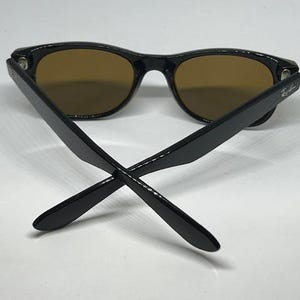Puede incluir: Gafas de sol con montura negra y lentes tintadas en marr&oacute;n. Las patillas est&aacute;n cruzadas en la parte delantera, mostrando el logotipo de Ray-Ban en el lado exterior de cada patilla. Las monturas tienen un acabado brillante y una forma cl&aacute;sica.