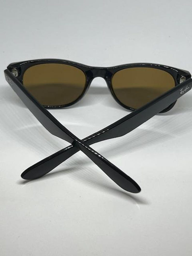 Puede incluir: Gafas de sol con montura negra y lentes tintadas en marr&oacute;n. Las patillas est&aacute;n cruzadas en la parte delantera, mostrando el logotipo de Ray-Ban en el lado exterior de cada patilla. Las monturas tienen un acabado brillante y una forma cl&aacute;sica.