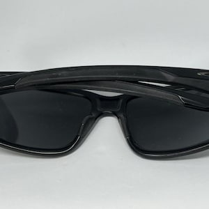 May include: Black sunglasses with dark, opaque lenses and a thick, glossy frame. The temples are folded inward over the front of the frames. A small, metallic logo emblem is visible on the outer hinge area of the temple on both sides.