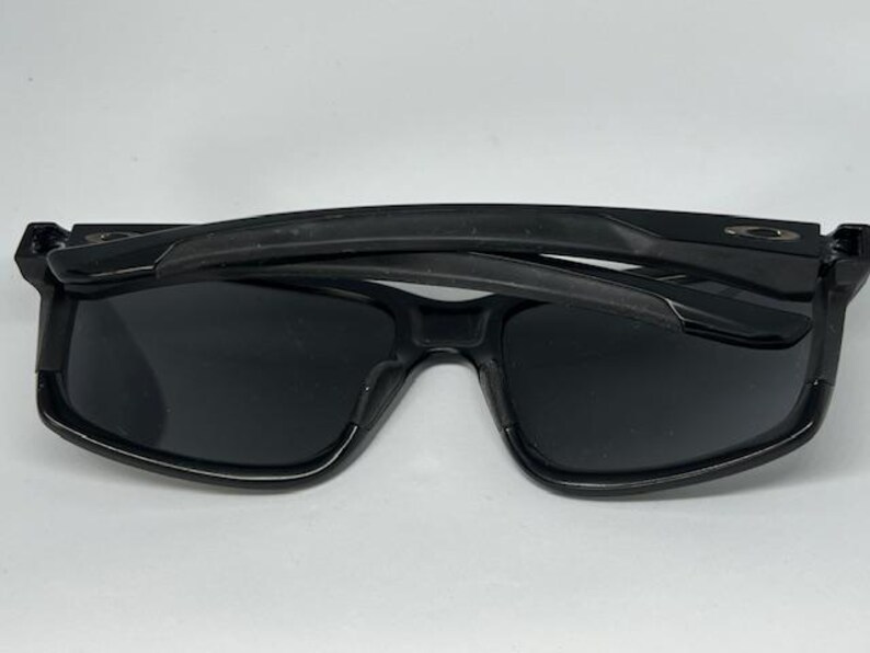 May include: Black sunglasses with dark, opaque lenses and a thick, glossy frame. The temples are folded inward over the front of the frames. A small, metallic logo emblem is visible on the outer hinge area of the temple on both sides.