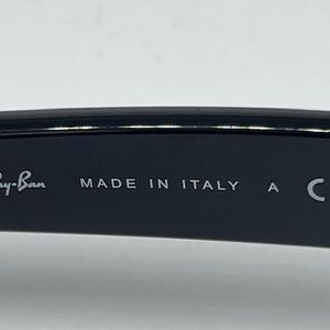 Puede incluir: El interior de una patilla de gafas negra con el texto impreso en blanco Ray-Ban, MADE IN ITALY, la letra A y la marca CE. La superficie es lisa y oscura, mostrando claramente la marca y los detalles de fabricaci&oacute;n en la montura.