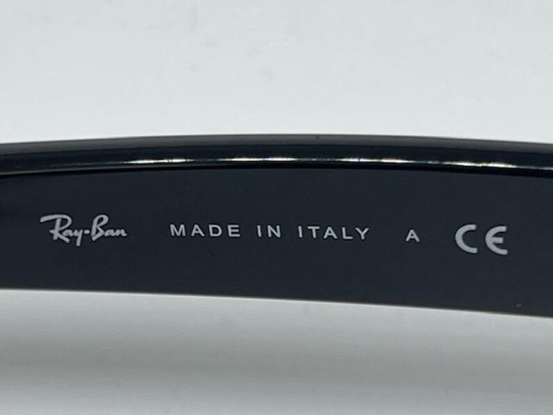 Puede incluir: El interior de una patilla de gafas negra con el texto impreso en blanco Ray-Ban, MADE IN ITALY, la letra A y la marca CE. La superficie es lisa y oscura, mostrando claramente la marca y los detalles de fabricaci&oacute;n en la montura.