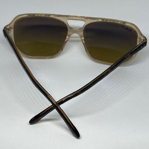 May include: Aviator sunglasses featuring a translucent beige frame with a double bridge design. The lenses have a gradient brown tint, and the temples are crafted from a dark brown tortoiseshell pattern material with the Ray-Ban logo visible.