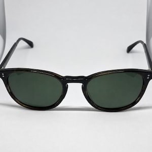 May include: Tortoiseshell-patterned sunglasses with dark green lenses. The frames feature a rounded shape with two small silver-toned metal rivets on each side of the front rim. The thin temples extend outward, showcasing the dark brown and amber mottled pattern.