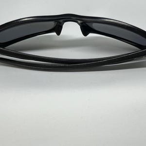 May include: A pair of black sunglasses with a curved, wrap-around frame design and dark, tinted lenses. The temples are folded inward, showcasing the sleek, solid black plastic construction of the eyewear from a top-down perspective.