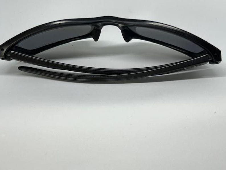 May include: A pair of black sunglasses with a curved, wrap-around frame design and dark, tinted lenses. The temples are folded inward, showcasing the sleek, solid black plastic construction of the eyewear from a top-down perspective.