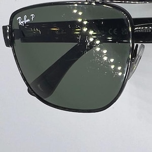 May include: Black metal frame sunglasses with dark green polarized lenses. The upper left lens features the white Ray-Ban P logo. The temple arm displays the CE mark and the word POLARIZED. The design includes a double bridge across the top.