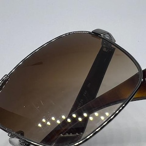 Pode incluir: &Oacute;culos de sol com arma&ccedil;&atilde;o met&aacute;lica escura e lentes castanhas. A haste interior apresenta o texto impresso 490 15 140 3N. O design inclui almofadas nasais transparentes e hastes castanhas escuras com padr&atilde;o de tartaruga para um visual cl&aacute;ssico.