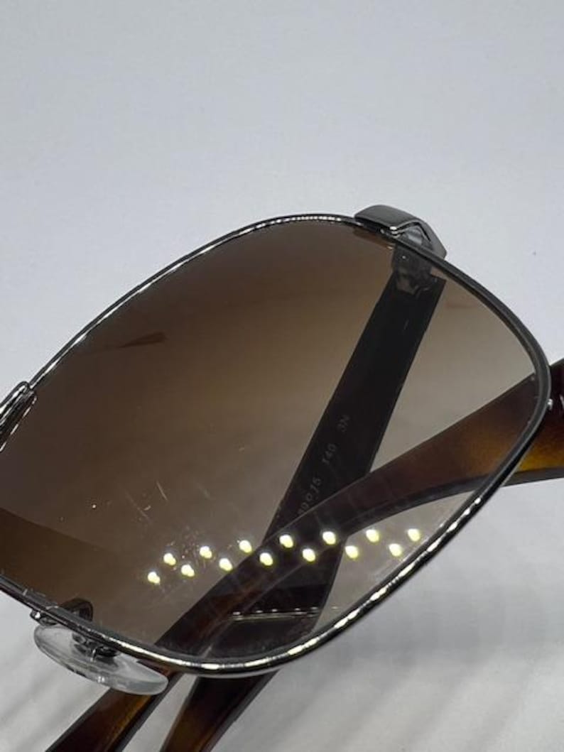 Pode incluir: &Oacute;culos de sol com arma&ccedil;&atilde;o met&aacute;lica escura e lentes castanhas. A haste interior apresenta o texto impresso 490 15 140 3N. O design inclui almofadas nasais transparentes e hastes castanhas escuras com padr&atilde;o de tartaruga para um visual cl&aacute;ssico.
